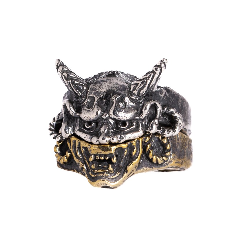 Puede incluir: Un anillo de plata y oro con una m&aacute;scara oni tridimensional detallada con cuernos. La m&aacute;scara tiene una expresi&oacute;n feroz con dientes y ojos afilados.