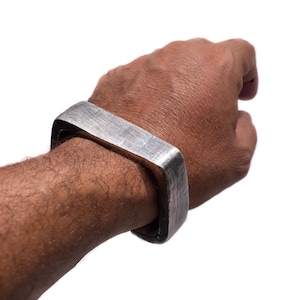 Könnte beinhalten: Ein silberfarbenes, quadratisch geformtes Armband wird am Handgelenk getragen. Das Armband hat eine strukturierte Oberfläche und ein solides, industrielles Design. Der Hintergrund ist schlicht weiß.