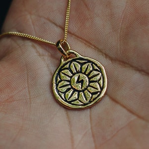 May include: A gold necklace with a round pendant featuring a stylized sun with a lightning bolt in the center. The pendant has a detailed design with black accents.