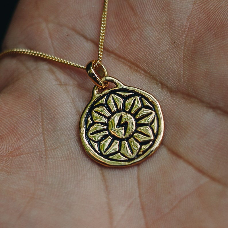 May include: A gold necklace with a round pendant featuring a stylized sun with a lightning bolt in the center. The pendant has a detailed design with black accents.