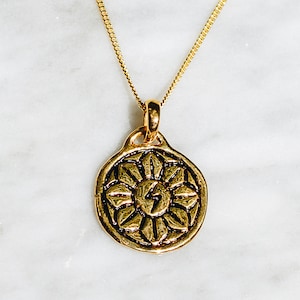 May include: Gold necklace with a round pendant featuring a stylized sun with a lightning bolt in the center. The pendant has a detailed design with a floral pattern.