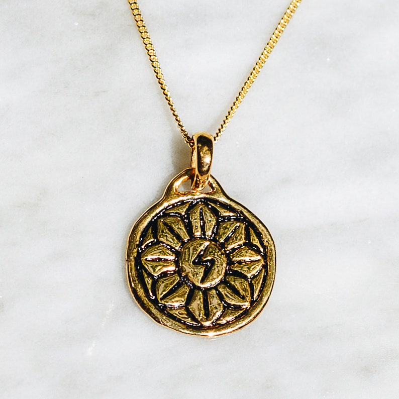 May include: Gold necklace with a round pendant featuring a stylized sun with a lightning bolt in the center. The pendant has a detailed design with a floral pattern.