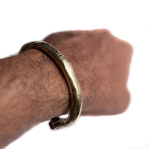 Puede incluir: Un brazalete de puño abierto de color dorado con una superficie texturizada. El brazalete se lleva en la muñeca de una persona, con el brazo y la mano visibles sobre un fondo blanco. La joya tiene un aspecto rústico y artesanal.
