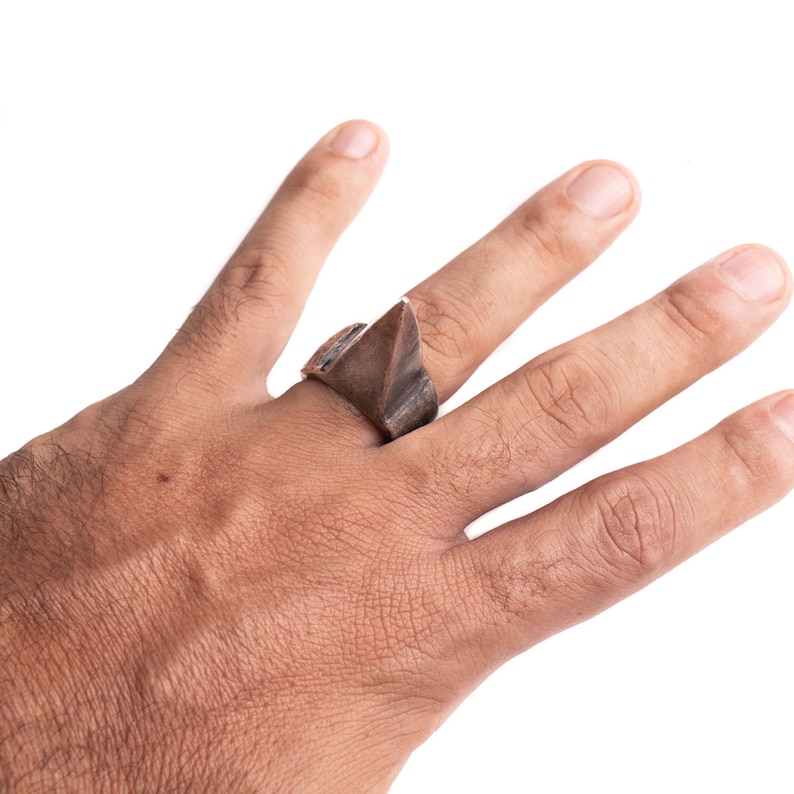 May include: A silver ring with a geometric, triangular design. The ring is worn on the middle finger of a hand.