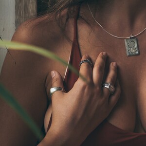 May include: A close-up of a person's chest with a silver necklace and pendant. The pendant is a rectangular shape with a design of a woman's face. The person is wearing a red top and has their hand on their chest. They are wearing several silver rings on their fingers.