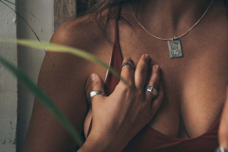May include: A close-up of a person's chest with a silver necklace and pendant. The pendant is a rectangular shape with a design of a woman's face. The person is wearing a red top and has their hand on their chest. They are wearing several silver rings on their fingers.