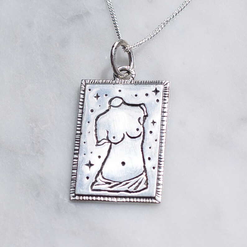 May include: Silver necklace with a rectangular pendant featuring a stylized outline of a female torso with a star pattern background.