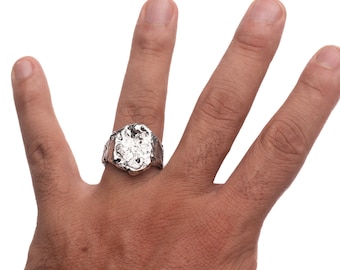May include: A silver ring with a textured, irregular oval-shaped top. The ring is worn on the index finger of a hand.