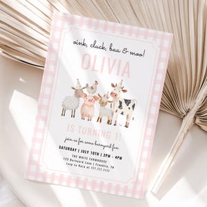 Pode incluir: Um convite com borda xadrez rosa com o texto "oink, cluck, baa & moo! OLIVIA IS TURNING 1!" apresenta ilustrações de animais de fazenda. O convite está em um prato branco com uma folha de palmeira seca.