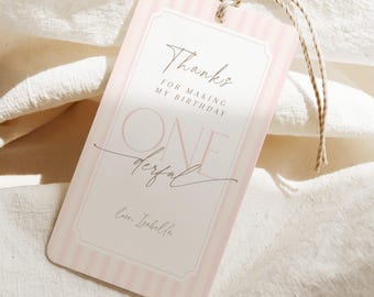 Puede incluir: Una etiqueta de regalo a rayas rosas y blancas con las palabras "Thanks for making my birthday" y "One derful" impresas. La etiqueta tiene un borde decorativo y una cuerda para sujetarla a un regalo. La etiqueta está sobre un fondo de tela blanca.