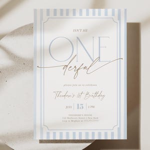 Puede incluir: Una tarjeta de invitación a rayas azul claro y blanco con el texto "ISN'T HE ONE derful" y "Theodore's 1st Birthday". La tarjeta está sobre una superficie circular blanca con una sombra de hoja.