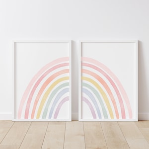 May include: Two framed prints of pastel watercolour rainbows, each with six arches in shades of pink, orange, yellow, green, blue, and purple. The prints are hung on a white wall.