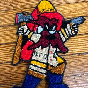 May include: A red, yellow, and blue felt patch of a cartoon firefighter holding an axe and pointing a gun. The firefighter is wearing a yellow helmet with the word "ARSON" on it. The firefighter has a red beard and the word "DEAD" is written on the front of the firefighter's jacket.