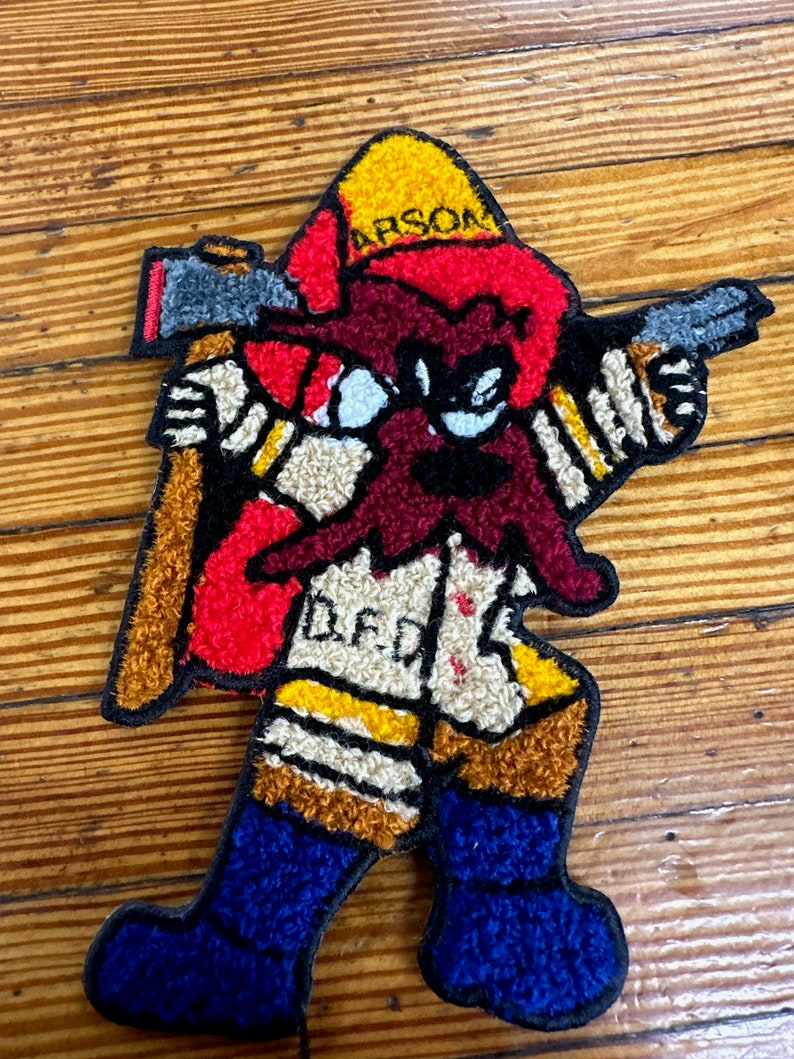 May include: A red, yellow, and blue felt patch of a cartoon firefighter holding an axe and pointing a gun. The firefighter is wearing a yellow helmet with the word "ARSON" on it. The firefighter has a red beard and the word "DEAD" is written on the front of the firefighter's jacket.