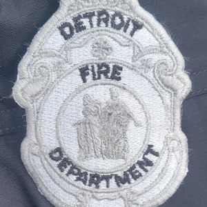 May include: A white embroidered patch with the words "DETROIT FIRE DEPARTMENT" in black lettering. The patch features a stylized image of a building with three figures standing in front of it.