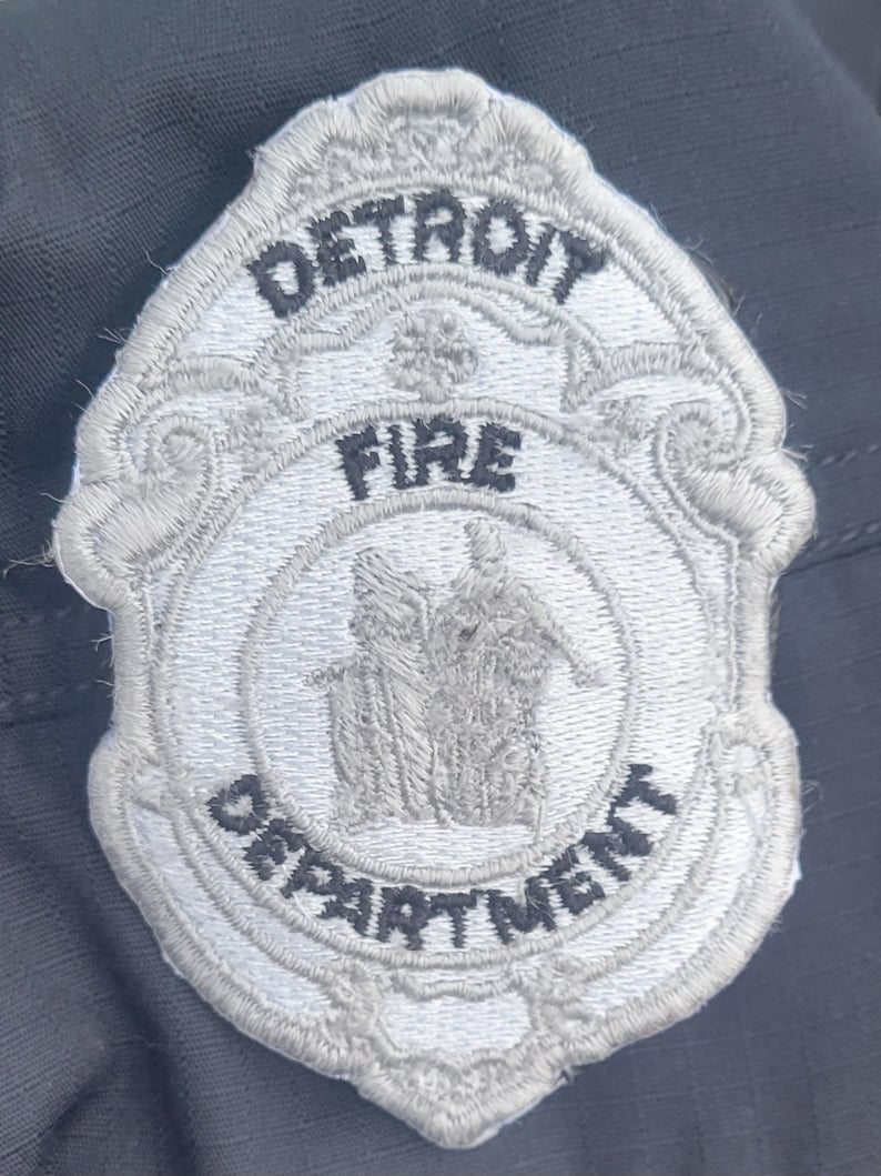 May include: A white embroidered patch with the words "DETROIT FIRE DEPARTMENT" in black lettering. The patch features a stylized image of a building with three figures standing in front of it.