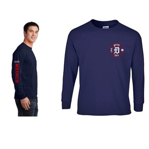 Pode incluir: Uma camiseta de manga comprida azul marinho com um círculo vermelho no peito. O círculo tem o texto "L.M.S. MOTOR CITY" e um "D" vermelho dentro. A manga esquerda tem a palavra "MEDICS" em vermelho.