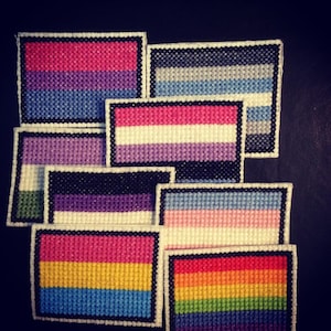 以下が含まれることがあります： 10種類の異なるプライドフラッグのコレクション。それぞれにユニークな配色とパターンが施されています。フラッグは布製で、縫い合わせられています。フラッグは暗い背景に表示されています。