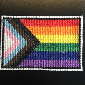 May include: A small, colorful embroidered patch featuring a rainbow flag with a transgender pride triangle in the corner. The patch is surrounded by a white border.