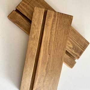 May include: Two wooden display stands with a groove for holding cards or signs. The stands are made of natural wood with a brown stain.