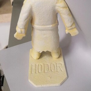 May include: A yellow painted figurine of a person wearing a robe, standing on a base with the word "HODOR" written on it.