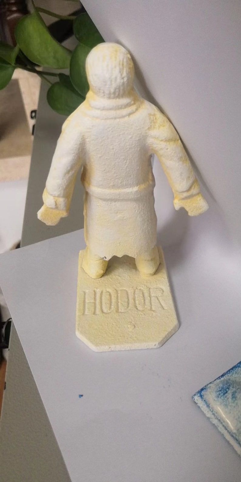May include: A yellow painted figurine of a person wearing a robe, standing on a base with the word "HODOR" written on it.