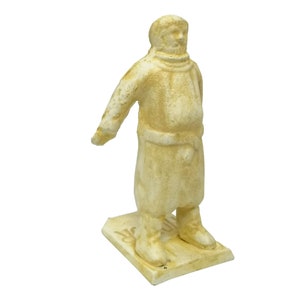 May include: A white painted figurine of a person wearing a long coat and boots. The figurine is standing on a square base with the word "Moscow" written on it.