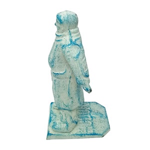 May include: A white cast iron figurine of a person wearing a blue and white winter coat standing on a base. The figurine is painted in a distressed white finish with blue accents.