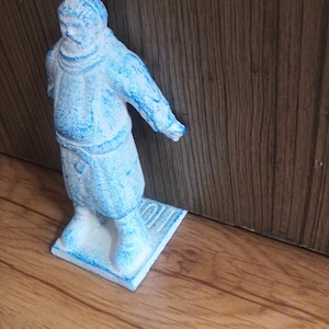 May include: A blue ceramic figurine of a person wearing a long robe. The figurine is standing on a small, rectangular base.
