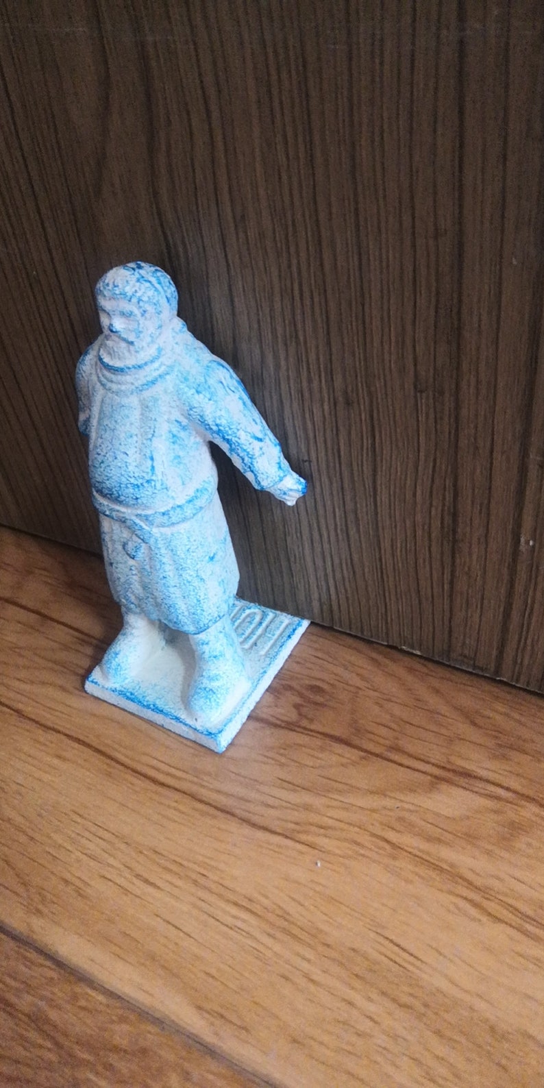 May include: A blue ceramic figurine of a person wearing a long robe. The figurine is standing on a small, rectangular base.