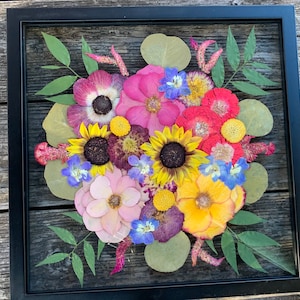 May include: A framed pressed flower arrangement with pink, red, yellow, and purple flowers. The flowers are arranged in a bouquet shape and are surrounded by green leaves.