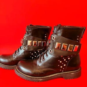 May include: Pair of black leather combat boots with silver studs and decorative straps. The boots have a lace-up design and a chunky sole. The straps are adorned with rectangular metal accents. The boots are set against a red background.