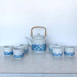 May include: A white ceramic tea set with a teapot and six cups. The teapot has a woven handle. The set is decorated with a blue wave pattern. The teapot has a spout and a lid. The cups and teapot are made of ceramic.