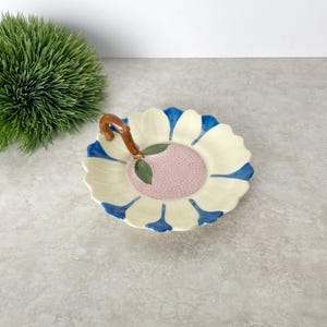 May include: A ceramic serving dish in the shape of a flower. The dish has a cream-colored base with blue petal tips, a pink center, and a brown handle. A green plant is in the background.