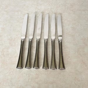 May include: Six silver butter knives with a decorative handle design. The blades are smooth and the handles have a flared shape with a beaded edge. The knives are arranged in a row on a light-colored surface.