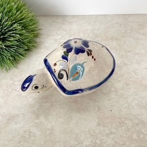 May include: A ceramic turtle figurine with a cream body and blue accents. The shell features blue floral designs and a blue border. The head and flippers are also blue, creating a decorative piece. A charming ornament for the home.