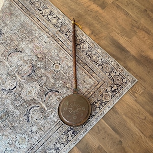 May include: A vintage copper and wood stove top kettle with a long wooden handle. The kettle has a decorative design on the front and is sitting on a patterned rug.