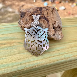 May include: A silver-toned owl pendant with a mother-of-pearl body and intricate wire detailing. The owl pendant is displayed on a piece of driftwood, set against a backdrop of green wooden planks.