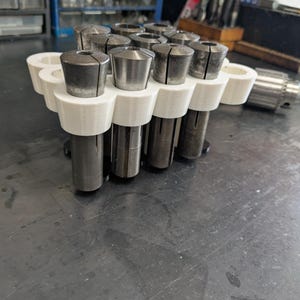 May include: A white 3D printed holder with 10 slots for storing metal collet chucks. The chucks are arranged in two rows of five. The holder is sitting on a gray metal surface.