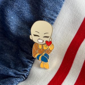May include: A gold-colored enamel pin featuring a cartoon character with a bald head, wearing a brown jacket, blue pants, and yellow shoes. The character is holding a red heart in its hand.