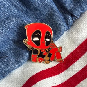 May include: A gold-colored enamel pin featuring a cartoon depiction of Deadpool, a superhero known for his red and black suit, sitting with his legs crossed. The pin has a detailed design with a red and black suit, white eyes, and a black mask.