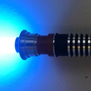 May include: A blue lightsaber with a silver and brown handle. The lightsaber is turned on and emitting a blue beam of light.