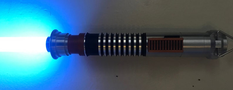 May include: A blue lightsaber with a silver and brown handle. The lightsaber is turned on and emitting a blue beam of light.