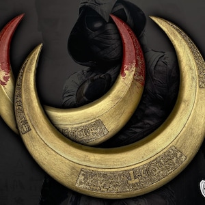 May include: Three golden crescent-shaped weapons with red tips, possibly props or replicas. The curved blades have intricate engraved patterns and a metallic sheen. The background features a shadowy figure, suggesting a theme of fantasy or action.
