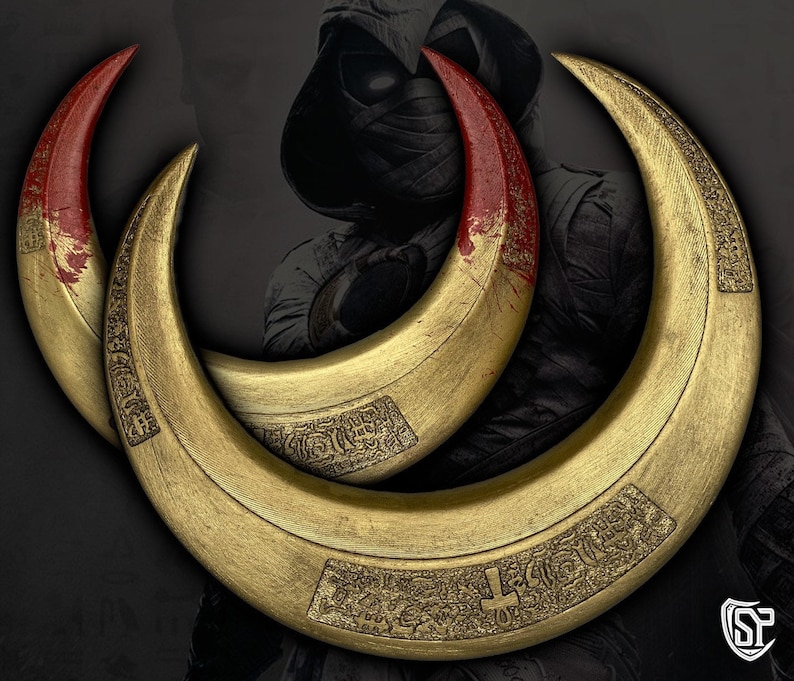 May include: Three golden crescent-shaped weapons with red tips, possibly props or replicas. The curved blades have intricate engraved patterns and a metallic sheen. The background features a shadowy figure, suggesting a theme of fantasy or action.