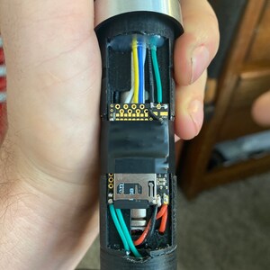 May include: A close-up of the inside of a lightsaber hilt, showing the wiring and electronics. The hilt is black with silver accents. The wiring is green, red, blue, yellow, and white. The electronics include a small circuit board and a battery.