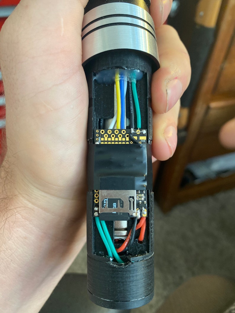 May include: A close-up of the inside of a lightsaber hilt, showing the wiring and electronics. The hilt is black with silver accents. The wiring is green, red, blue, yellow, and white. The electronics include a small circuit board and a battery.