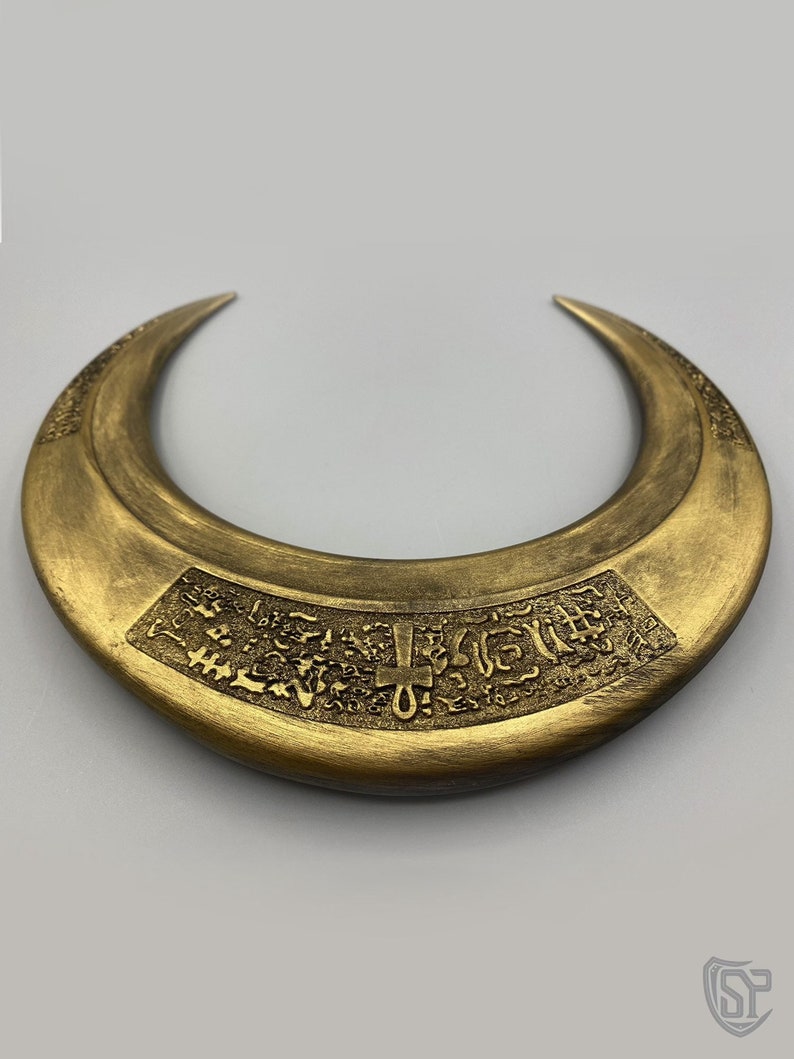 May include: A crescent-shaped, gold-colored necklace with engraved hieroglyphic-style symbols. The necklace has a wide band and pointed ends. The surface has a textured, metallic finish, and the design is centered on the front.