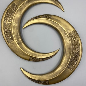 May include: Two bronze-colored, crescent-shaped objects with pointed ends. Each has an engraved design along the outer curve. The objects are arranged in a figure-eight pattern against a light gray background.
