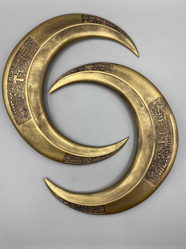 May include: Two bronze-colored, crescent-shaped objects with pointed ends. Each has an engraved design along the outer curve. The objects are arranged in a figure-eight pattern against a light gray background.